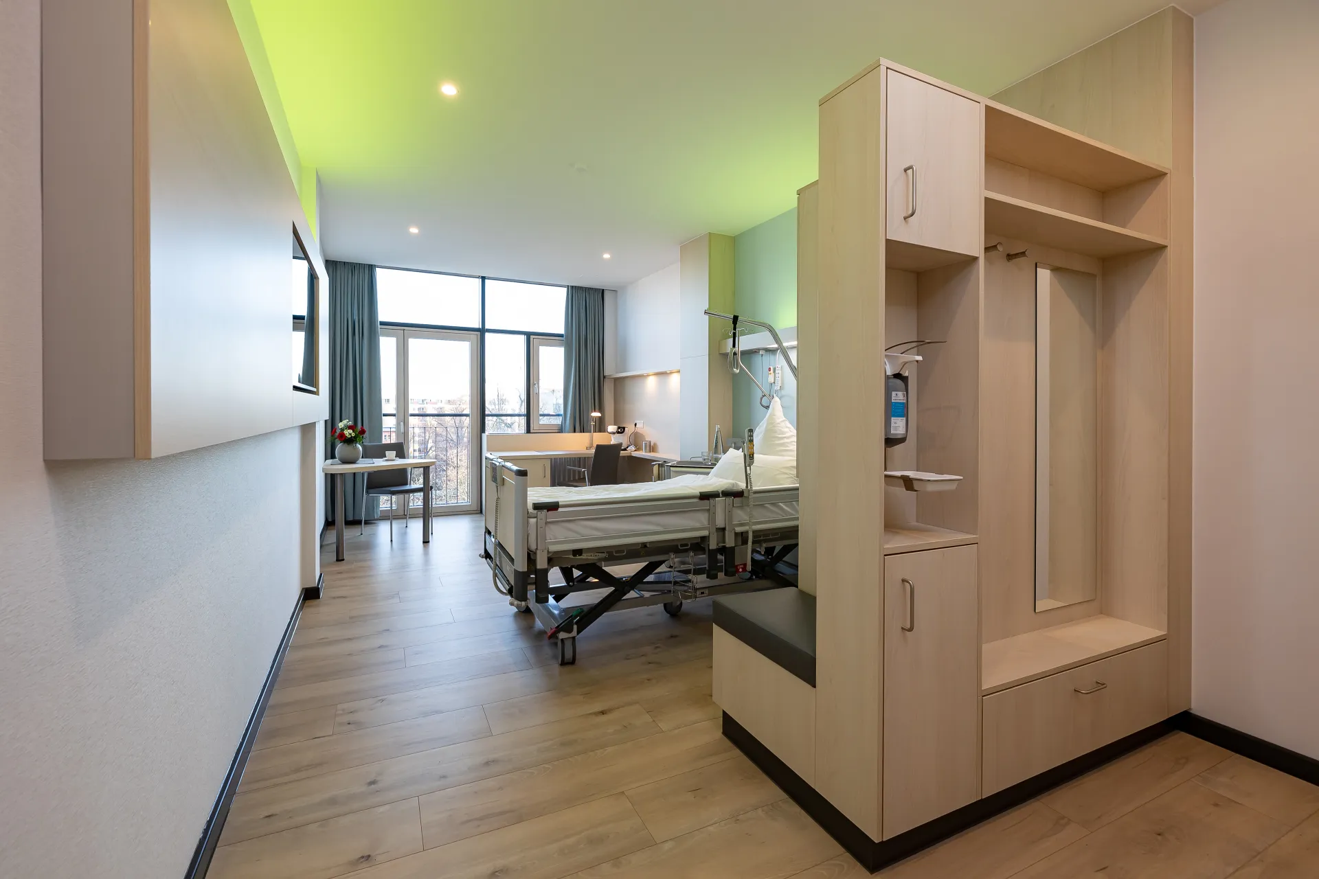 Modern eingerichtetes Patientenzimmer mit Pflegebett, Sitzbank, Schrank und großem Fenster mit Vorhängen