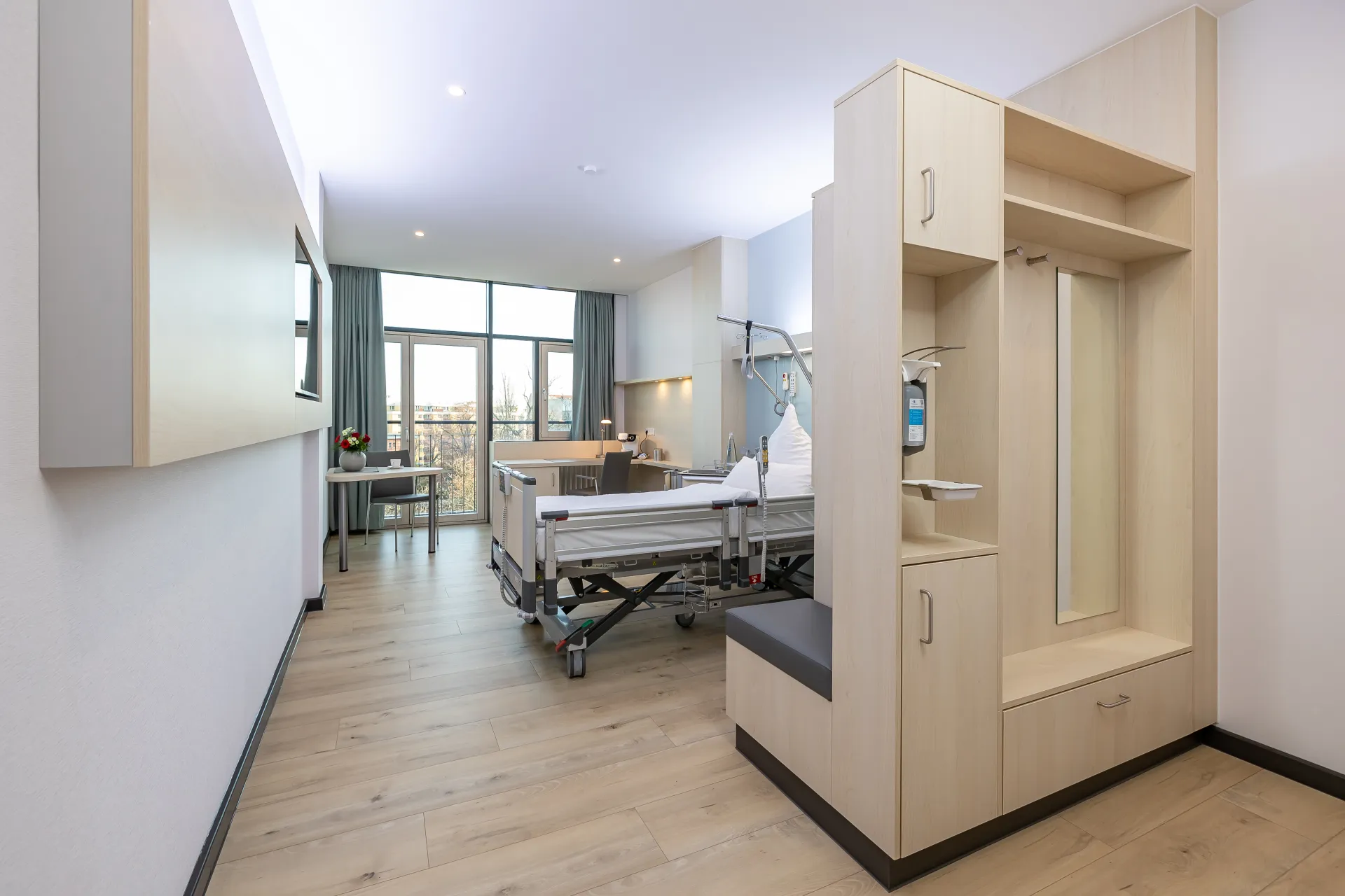 Helles Patientenzimmer mit Pflegebett, Sitzbank, Garderobe und großem Fenster mit Blick nach draußen
