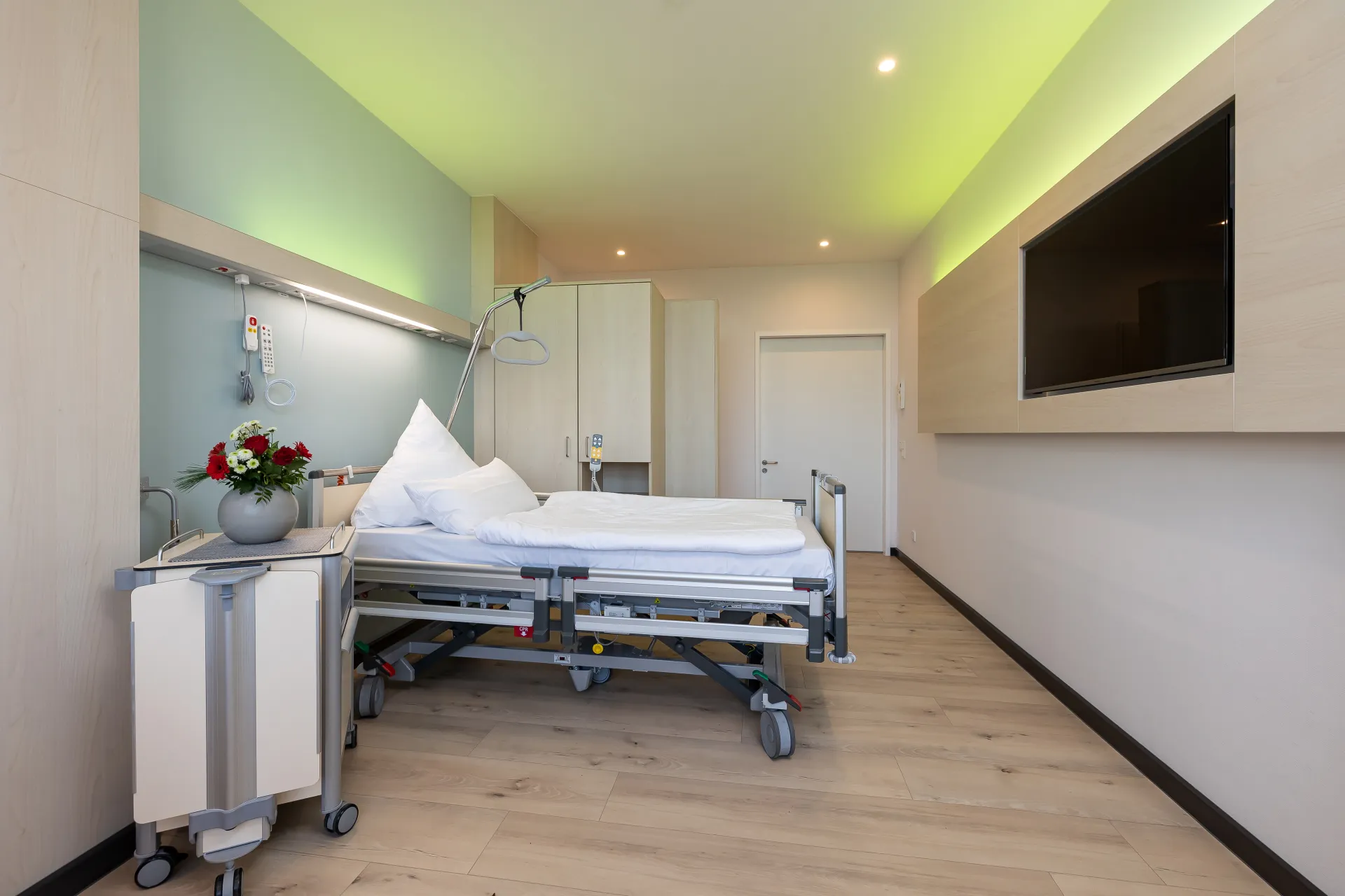 Krankenhauszimmer mit leerem Bett, Nachttisch mit Blumenvase und großem Wandfernseher