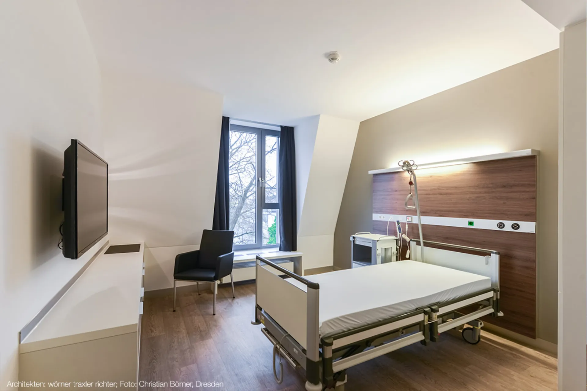 Ein schlicht eingerichtetes Patientenzimmer mit einem Krankenhausbett, einem schwarzen Stuhl, einem Fenster mit Vorhängen und einem Fernseher an der Wand