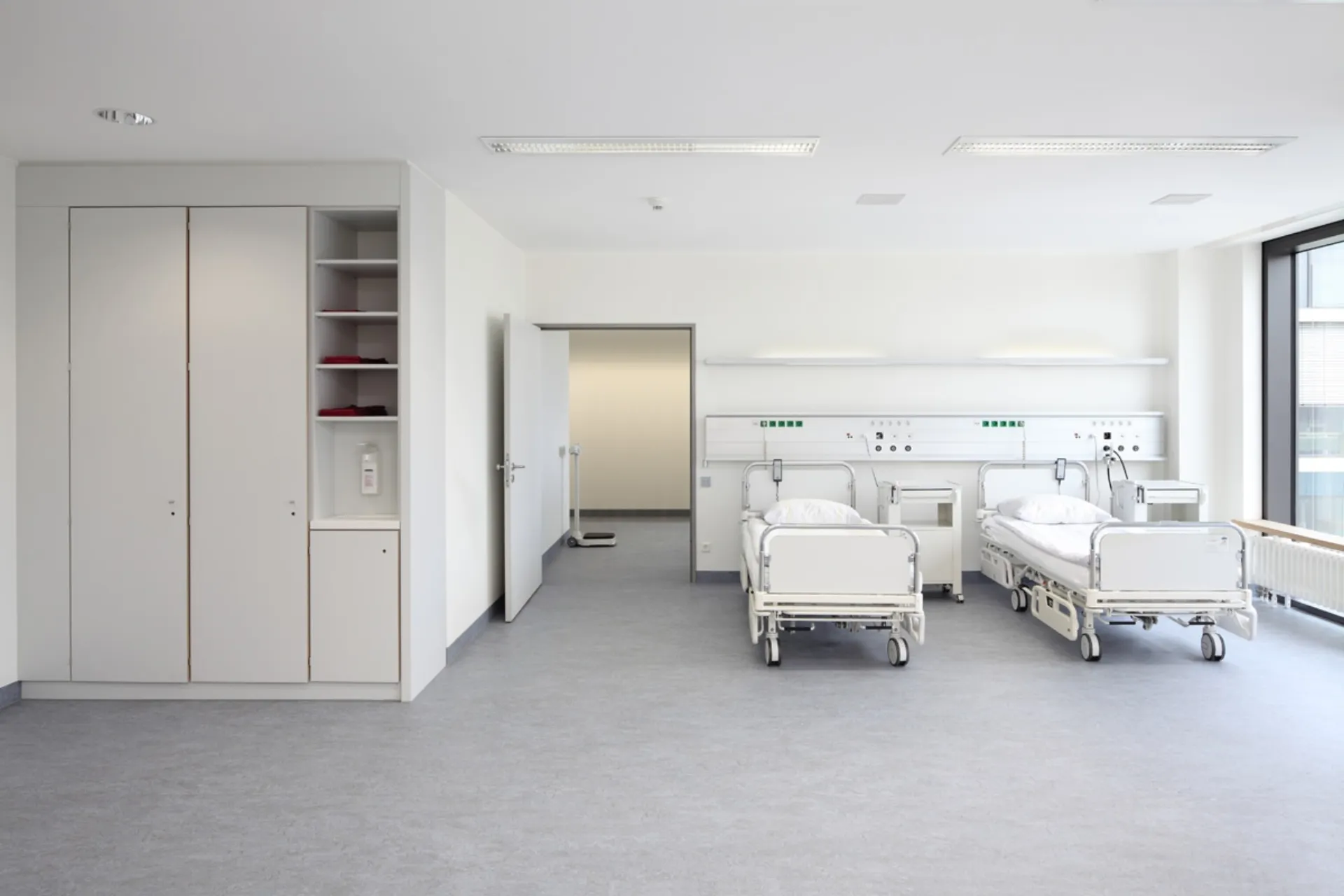 Zwei leere Krankenhausbetten mit weißen Bettwäsche in einem hellen Patientenzimmer mit Schrank und großem Fenster