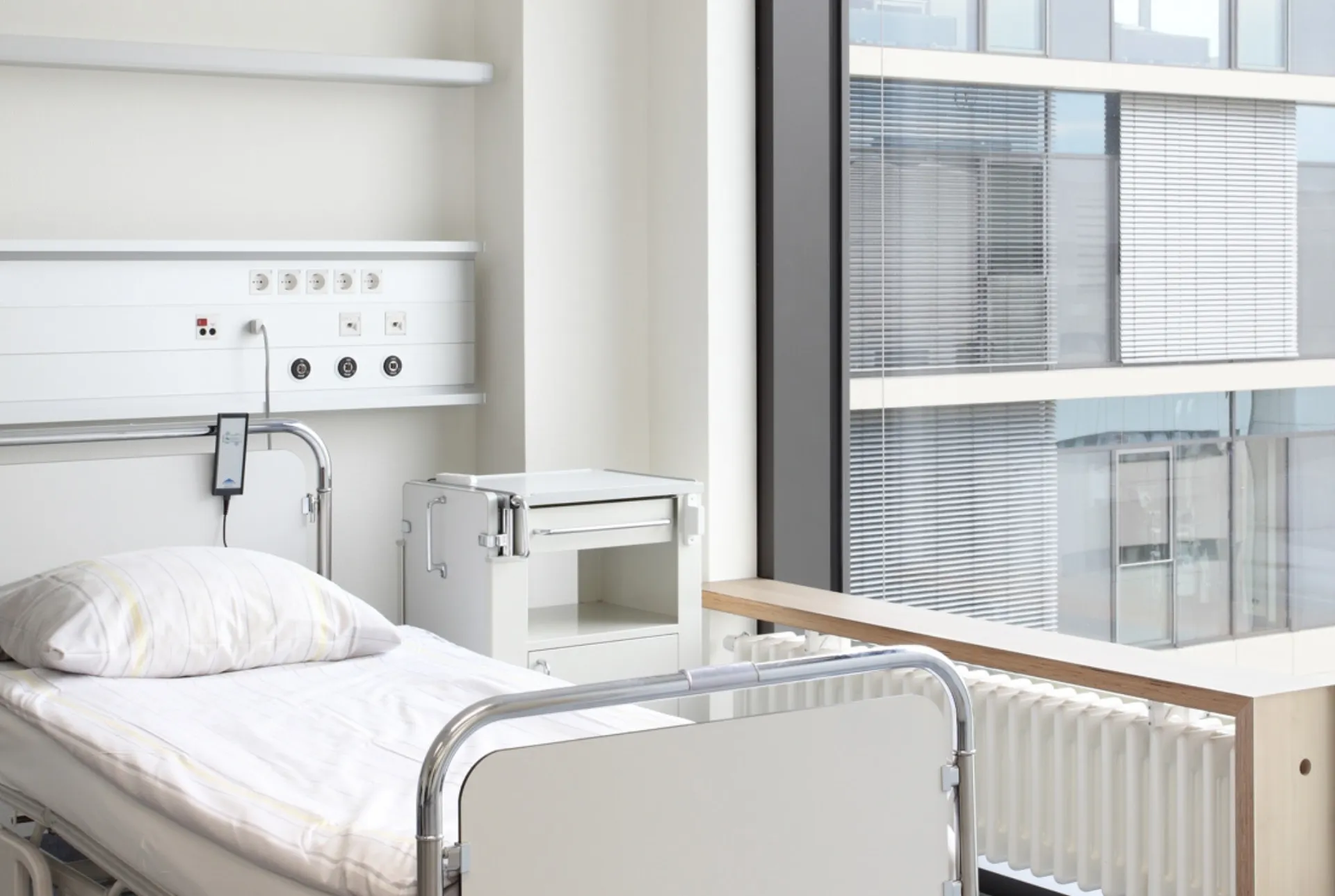Krankenhauszimmer mit leerem Bett, Nachttisch, medizinischer Wandkonsole und großem Fenster mit Blick auf ein Bürogebäude