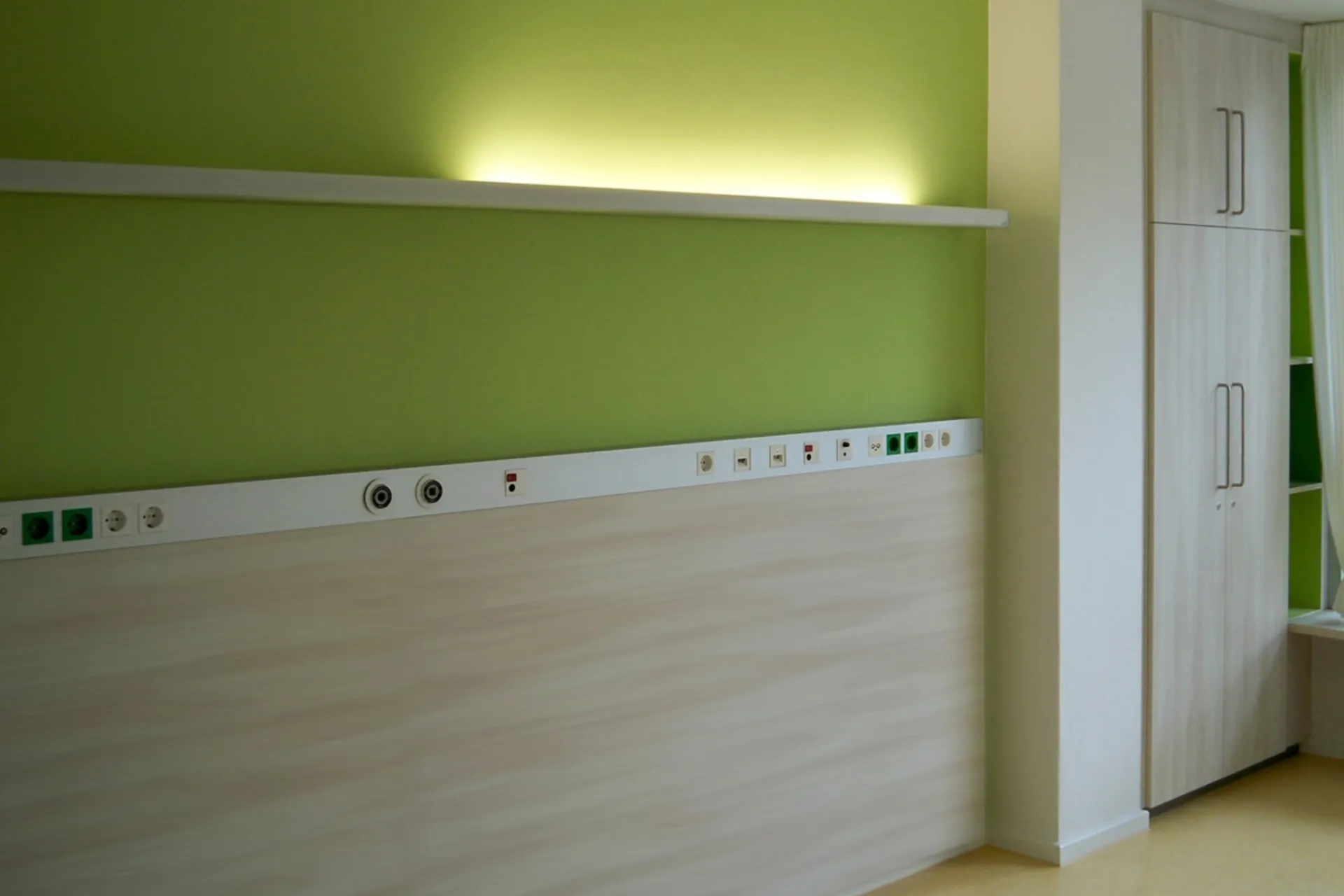 Wand mit mehreren Steckdosen und Anschlüssen unter einem beleuchteten weißen Regal in einem Raum mit grün gestrichener Wand und einem weißen Schrank
