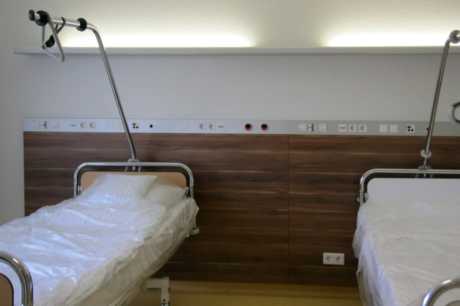 Zwei Krankenhausbetten mit weißen Bezügen und Aufrichthilfen vor einer Wand mit Holzverkleidung und medizinischen Anschlüssen