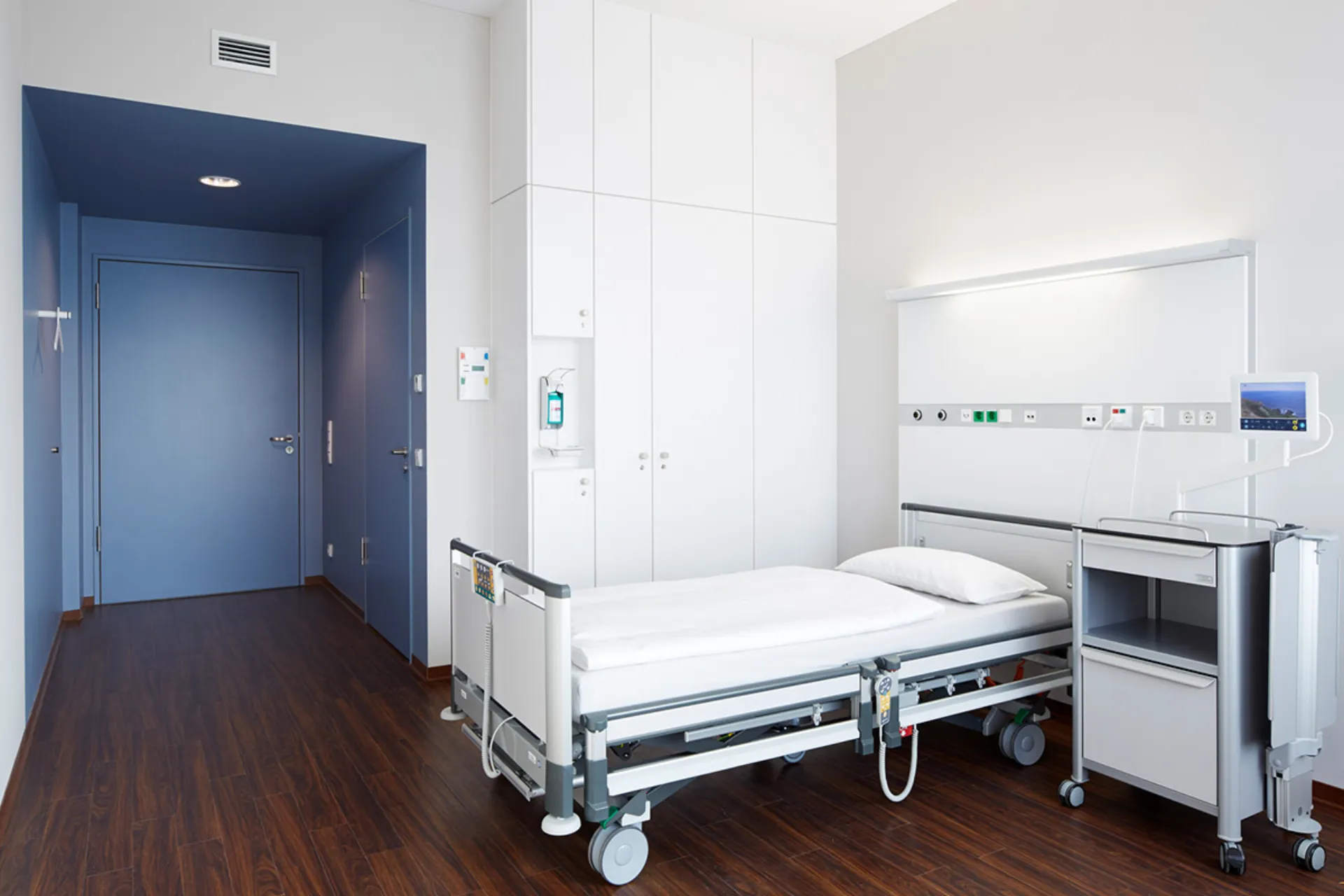 Krankenhauszimmer mit einem Bett, Nachttisch, weißen Schränken und blauer Tür in einem hellen Raum