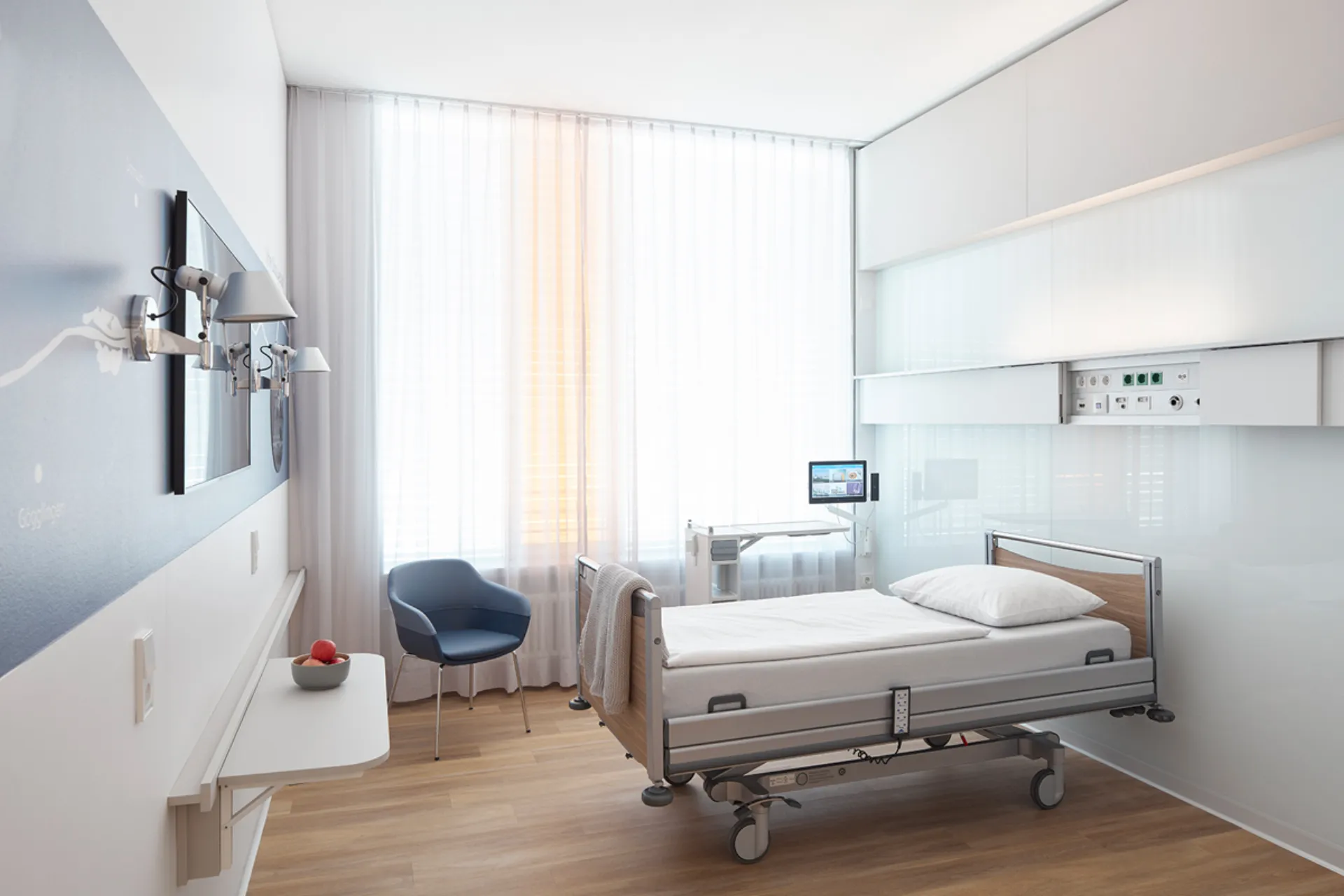 Modern eingerichtetes Einzelzimmer in einem Krankenhaus mit Pflegebett, blauem Stuhl und großem Fenster mit weißen Vorhängen, die Versorgungseinheit an der Wand ist aufgeschoben.
