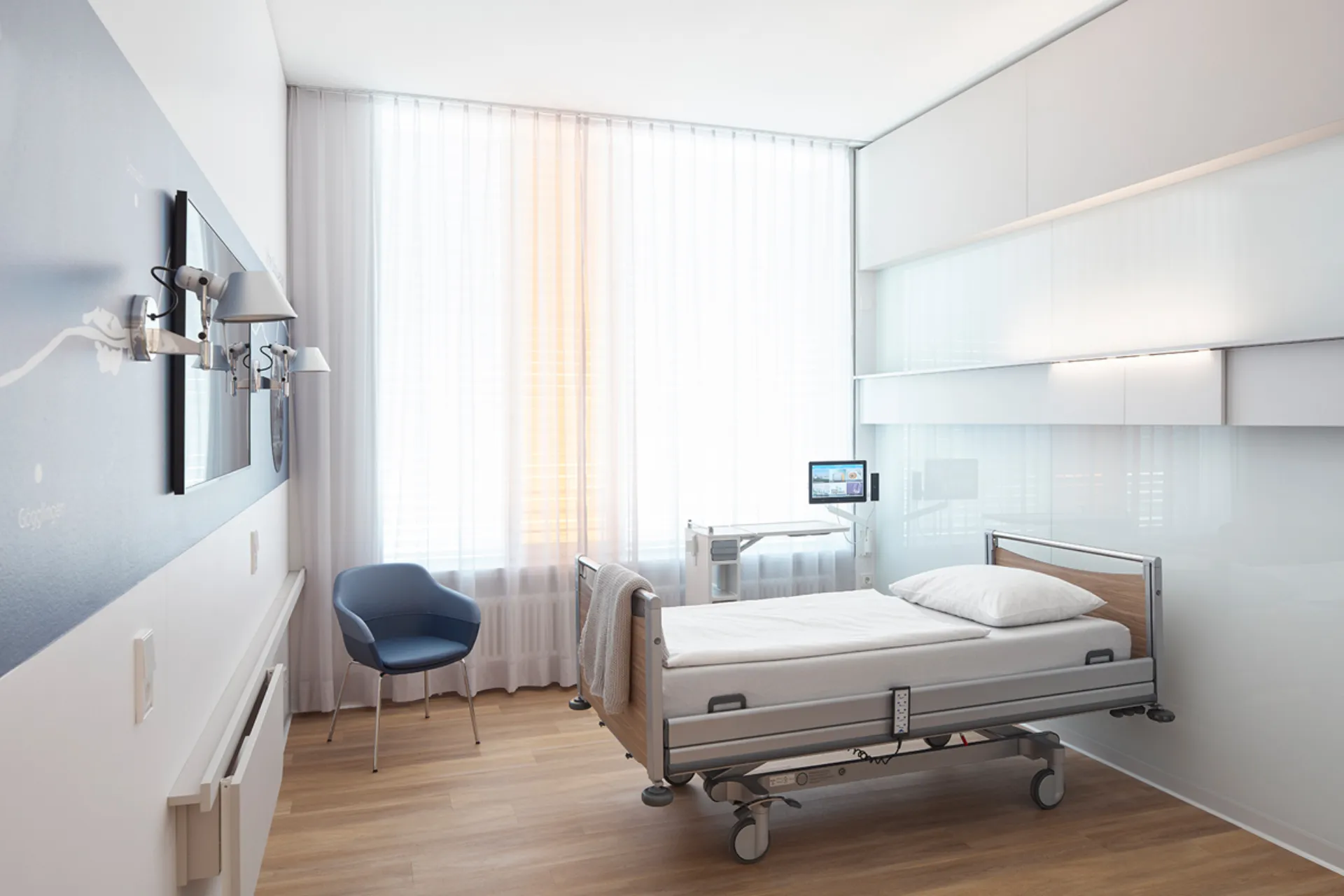 Modern eingerichtetes Einzelzimmer in einem Krankenhaus mit Pflegebett, blauem Stuhl und großem Fenster mit weißen Vorhängen
