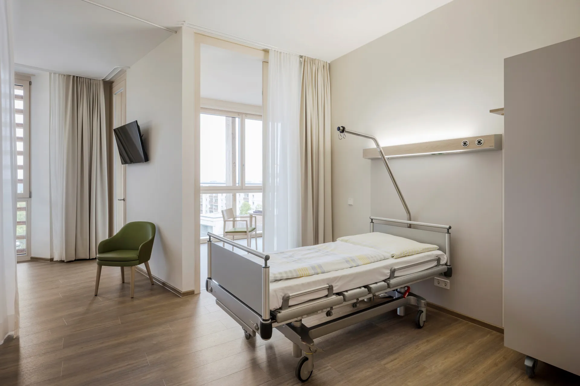 Krankenhauszimmer mit Pflegebett, grünem Stuhl, Fenster mit Blick auf Stadt und hellem Holzboden