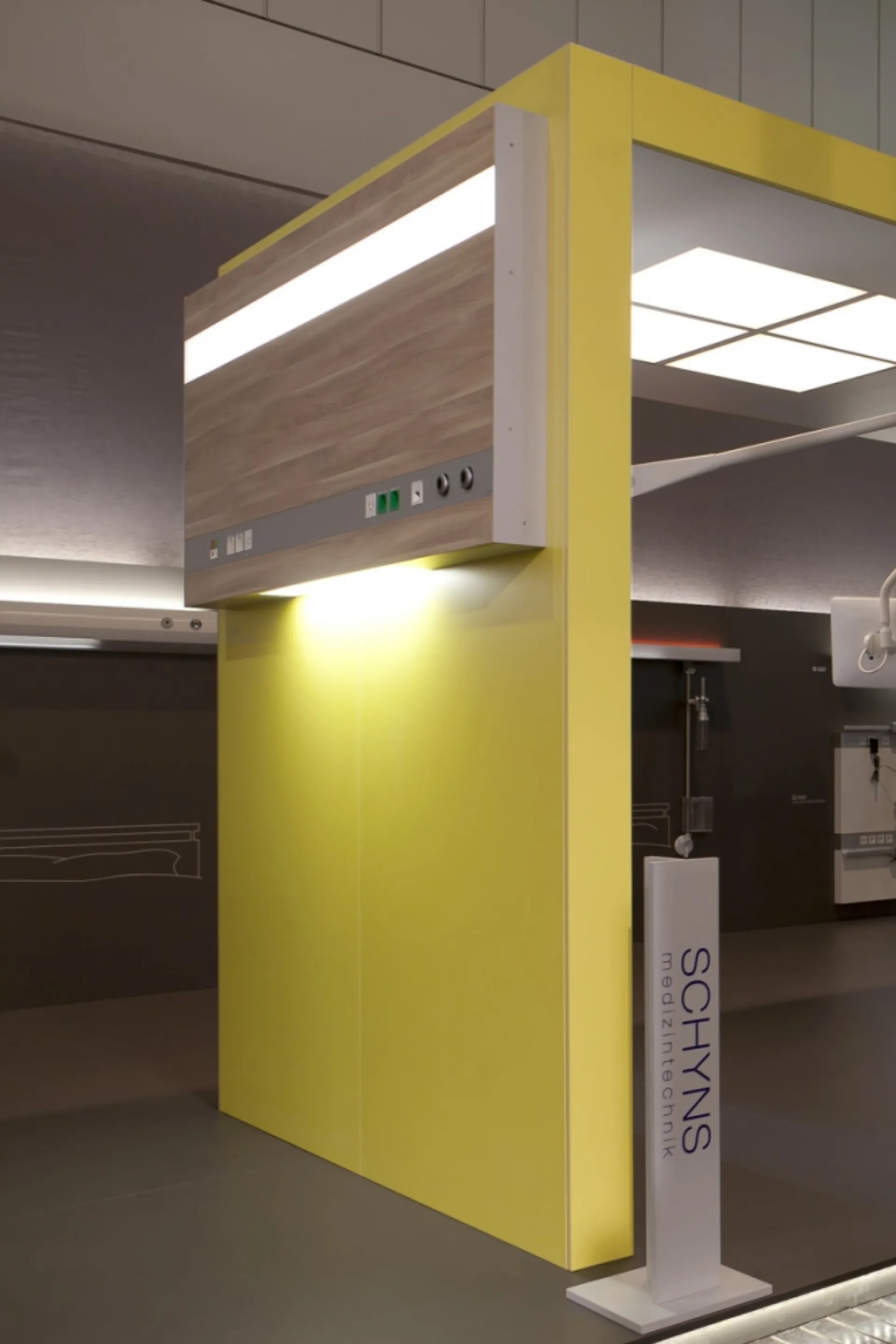 Gelb lackierte Wand mit Holzpaneel und integrierter Beleuchtung in einem Ausstellungsraum mit Stele 'SCHYNS Medizintechnik'
