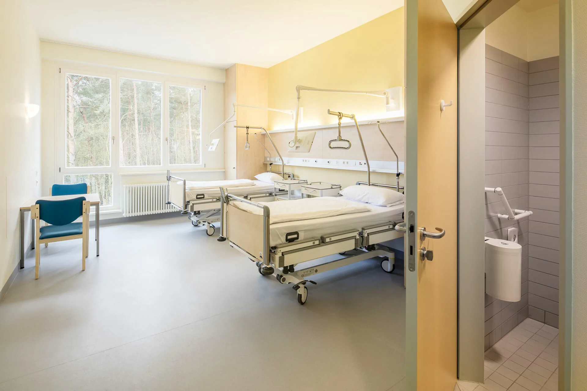 Zweibettzimmer mit gelber Akzentwand, indirekt beleuchteter Versorgungsleiste mit schwenkbaren Leselampen, Fenster mit Waldblick und offener Tür ins gefliestes Bad