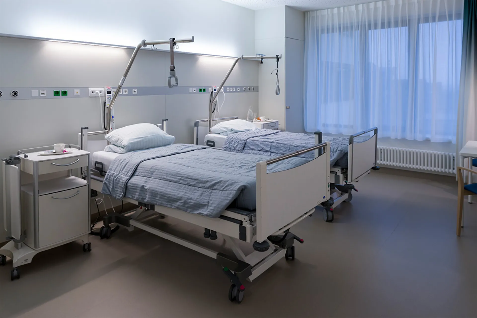 Zwei Krankenhausbetten mit grauen Decken in einem hellen Patientenzimmer mit großem Fenster und Vorhängen