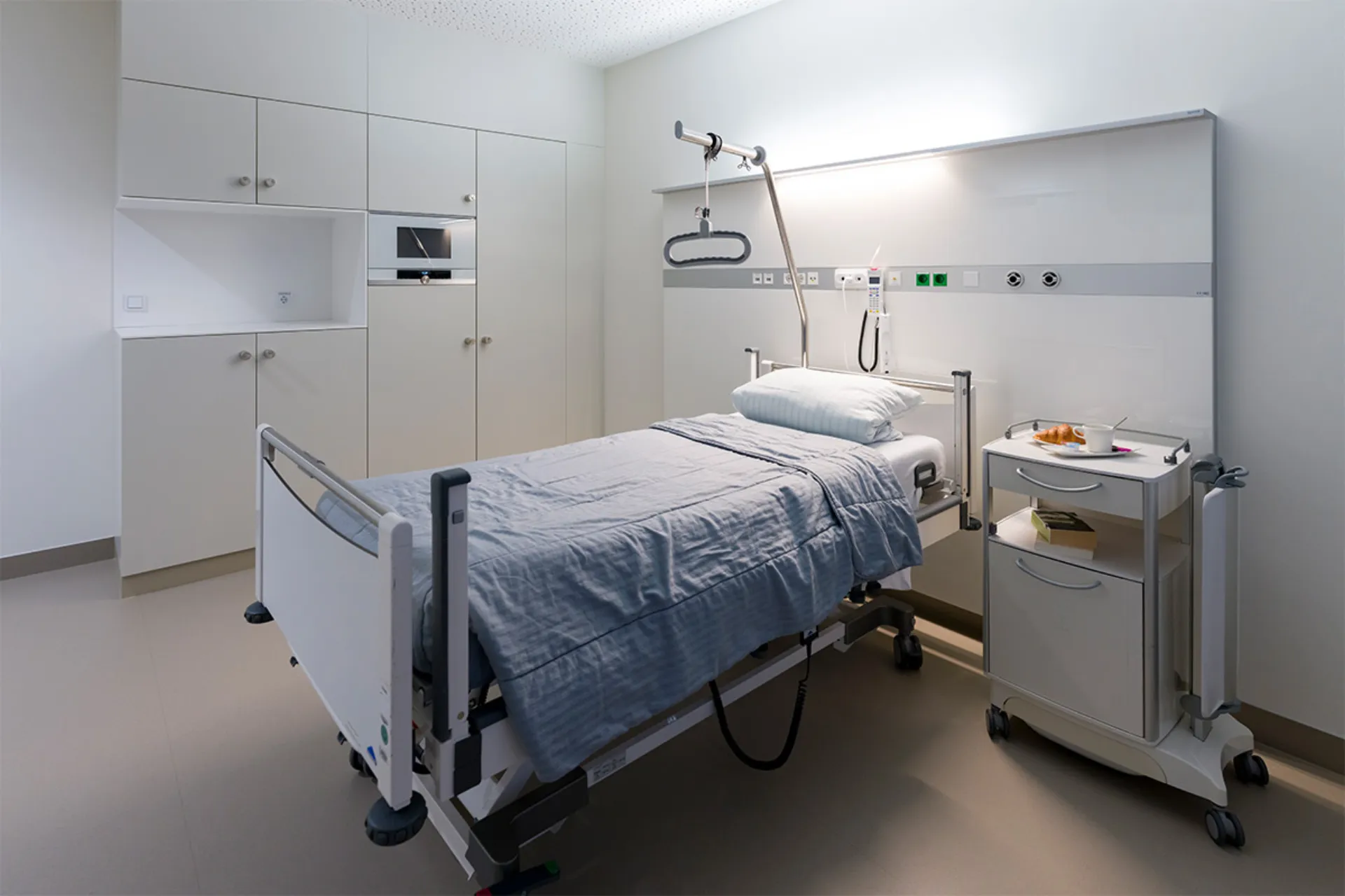 Krankenhauszimmer mit einem Bett, grauer Bettdecke, Nachttisch auf Rollen und weißen Schränken an der Wand