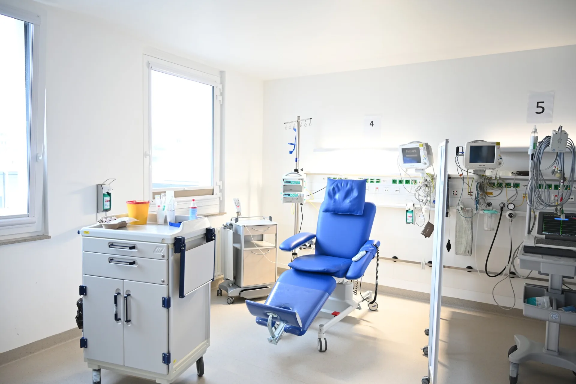 Dialysestation mit blauem Behandlungsstuhl, weißem Materialwagen, mehreren Patientenüberwachungsmonitoren an der Wand und großen Fenstern mit Tageslicht