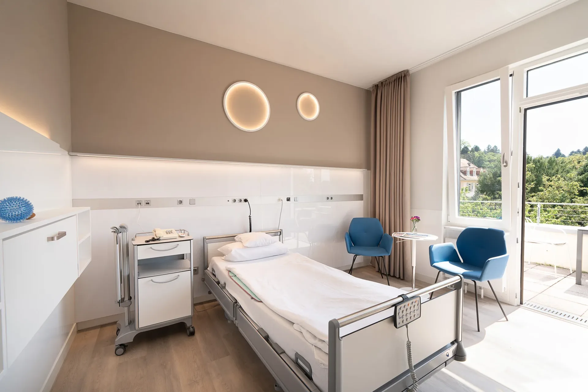 Modernes Patientenzimmer mit Pflegebett, Nachttisch auf Rollen, zwei runden Wandleuchten, zwei blauen Sesseln am Beistelltisch und Balkontür mit Ausblick ins Grüne