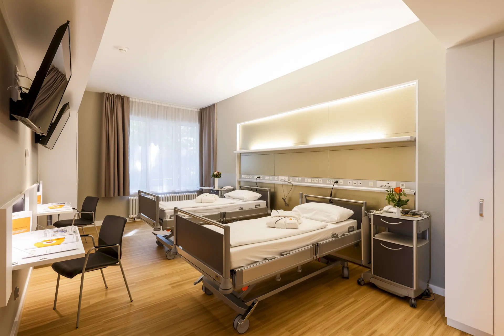 Patientenzimmer mit zwei Pflegebetten, indirekt beleuchteter Versorgungsleiste an der Rückwand, Schreibtisch mit Stuhl, Wandfernseher und Fenster mit Vorhang