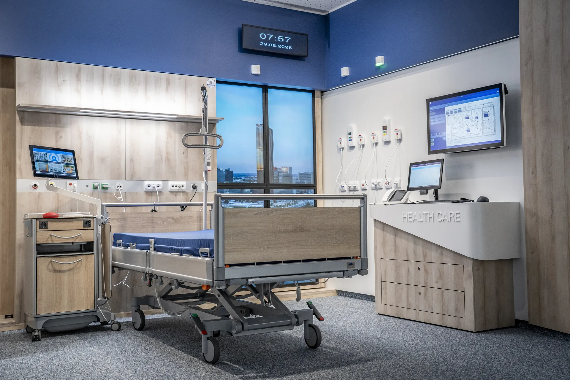 Modernes Intensivzimmer mit Pflegebett, blauer Akzentwand, digitaler Uhr 07:57, zwei Wandmonitoren, Pflegeplatz mit Healthcare-Schriftzug und Ausblick auf eine Skyline