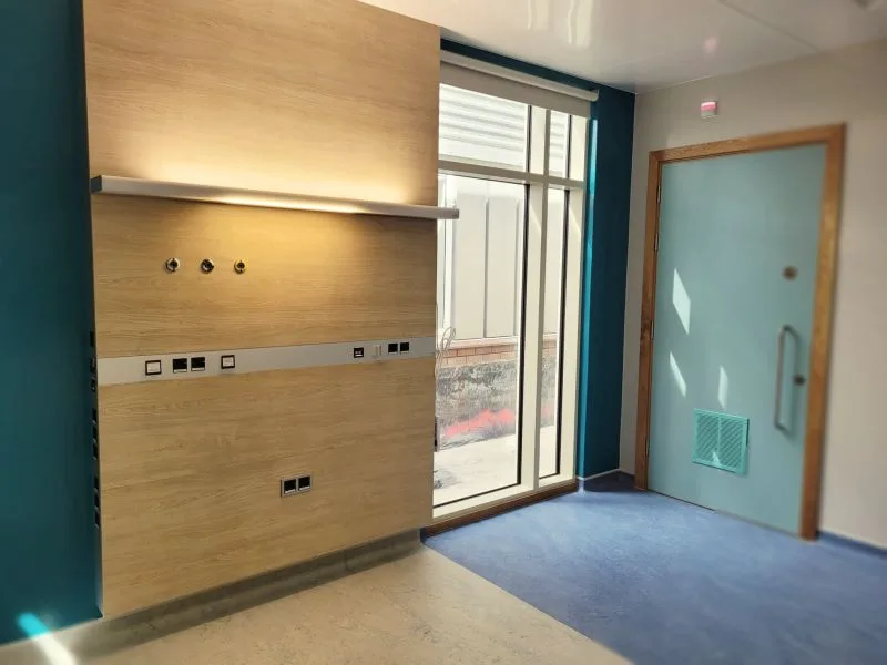 Wandverkleidung aus hellem Holz mit eingelassener Versorgungsleiste mit medizinischen Gas- und Stromanschlüssen vor tiefblauer Wand und Glastür