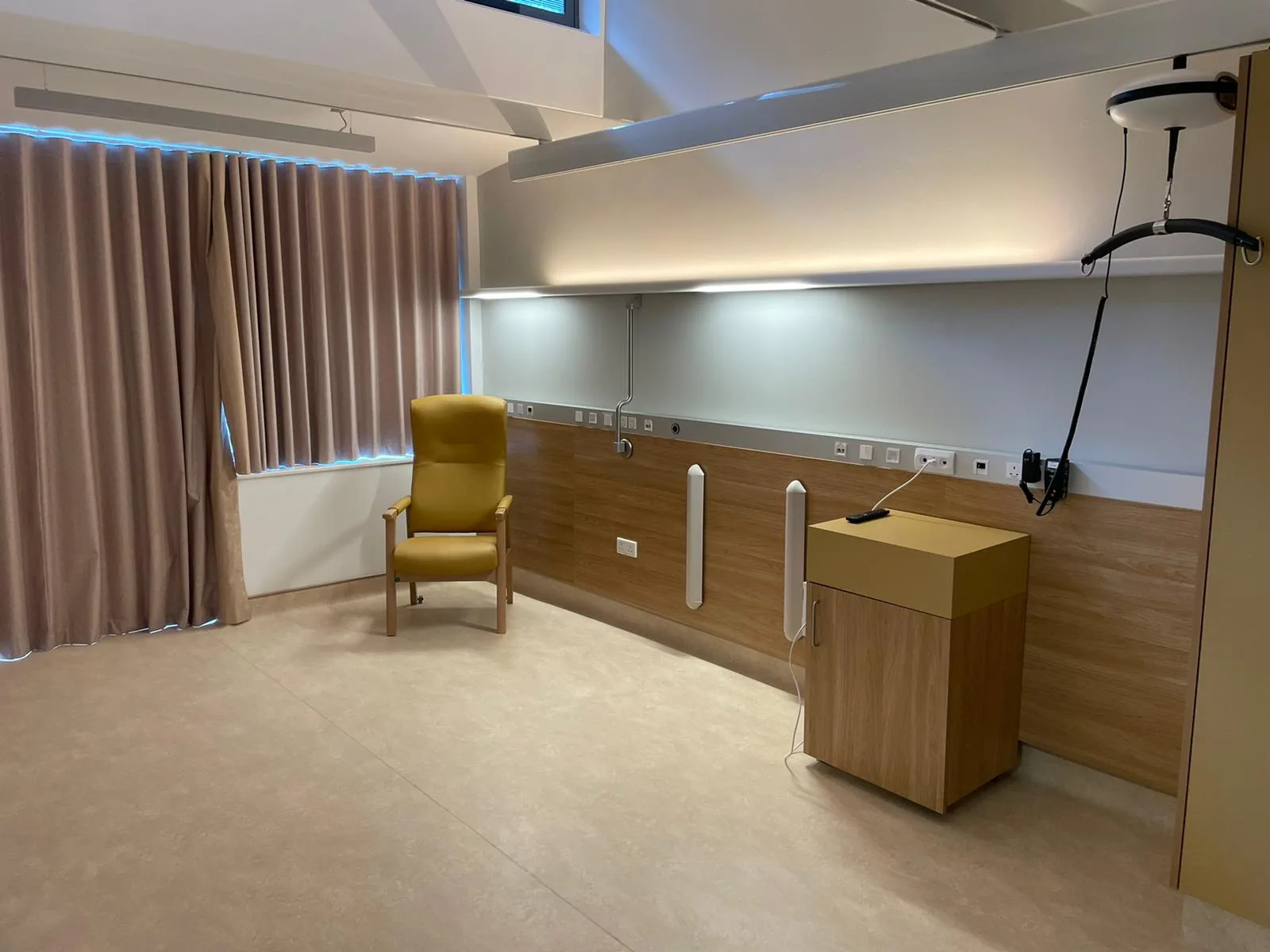 Patientenzimmer mit gelbem Ledersessel, Holzverkleidung mit medizinischen Gas- und Stromanschlüssen, indirekter Deckenbeleuchtung und Patientenlifter