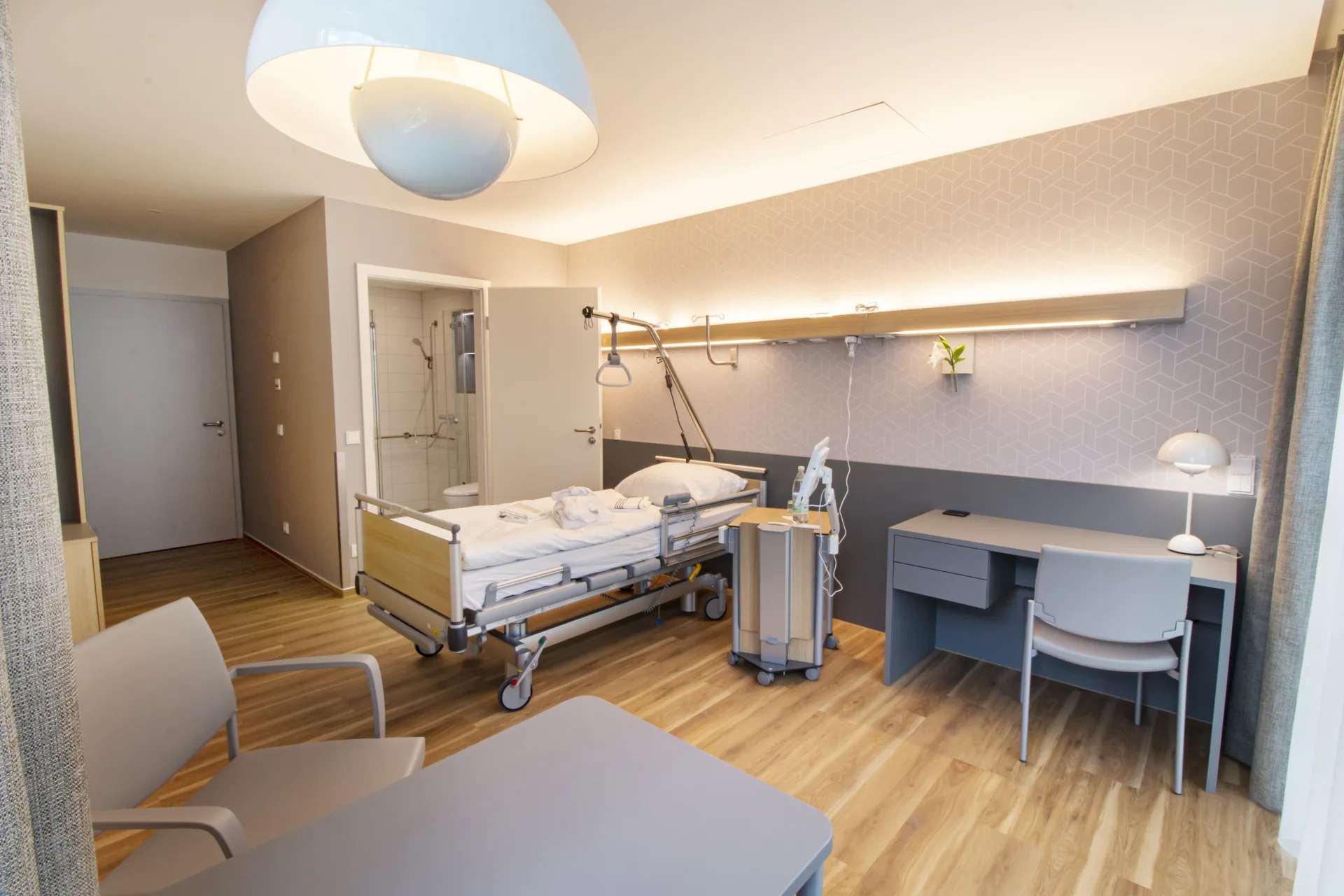 Modern eingerichtetes Patientenzimmer mit Klinikbett, Schreibtisch, Stühlen und eigenem Bad