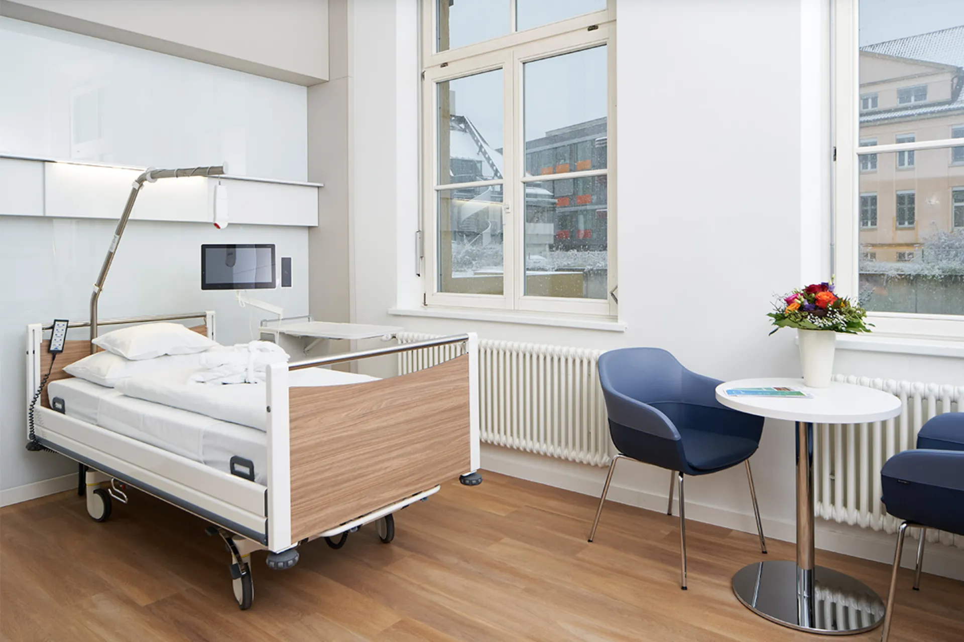 Krankenhauszimmer mit Pflegebett, zwei blauen Stühlen, rundem Tisch mit Blumenvase und großen Fenstern