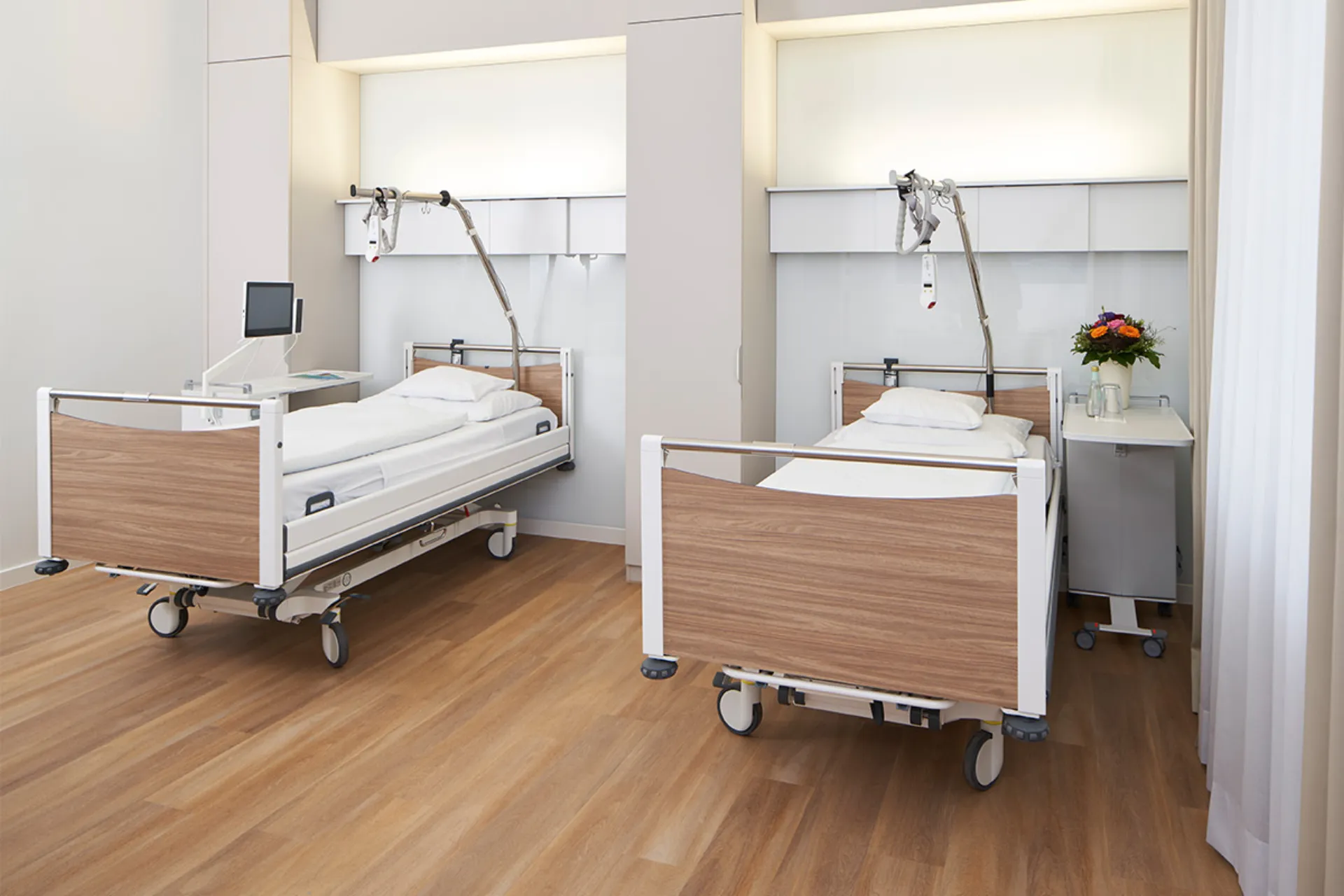 Zwei moderne Krankenhausbetten mit Holzverkleidung in einem hellen Patientenzimmer mit medizinischen Geräten und einem Blumenstrauß auf einem Rolltisch