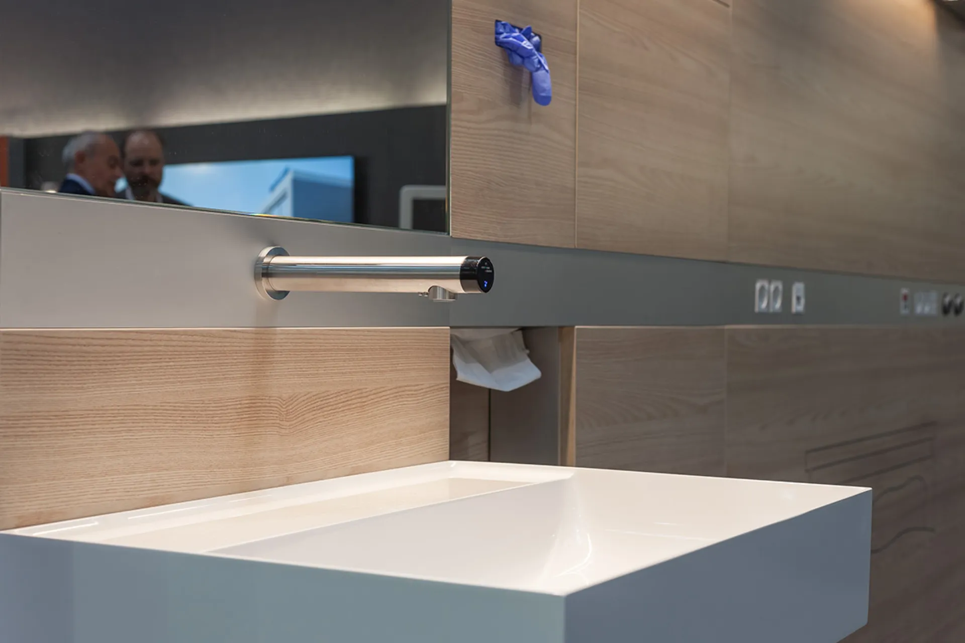 Moderner Waschtisch mit rechteckigem Becken, berührungsloser Armatur und Holzverkleidung an der Wand mit Spiegel und blauer Handschuhhalterung
